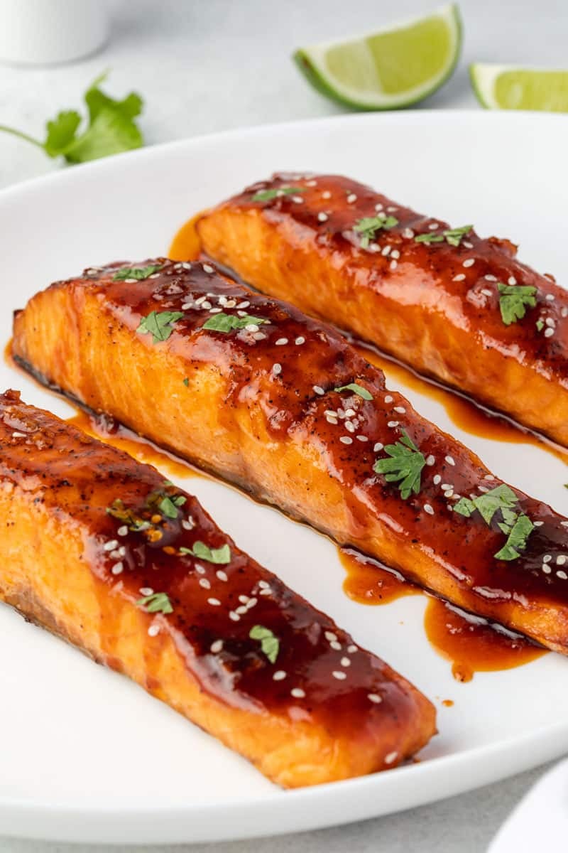 3 fillets of salmon on a white plate with honey sriracha sauce, sesame seeds, and chopped cilantro.
