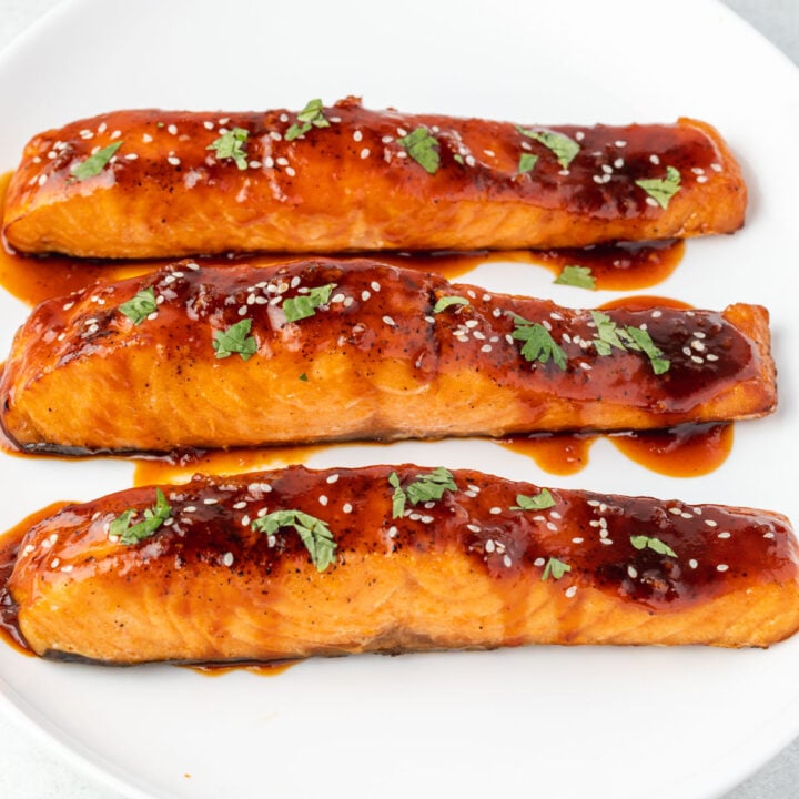 3 pieces of salmon in vertical rows on a plate, with honey sriracha sauce, sesame seeds, and chopped cilantro.
