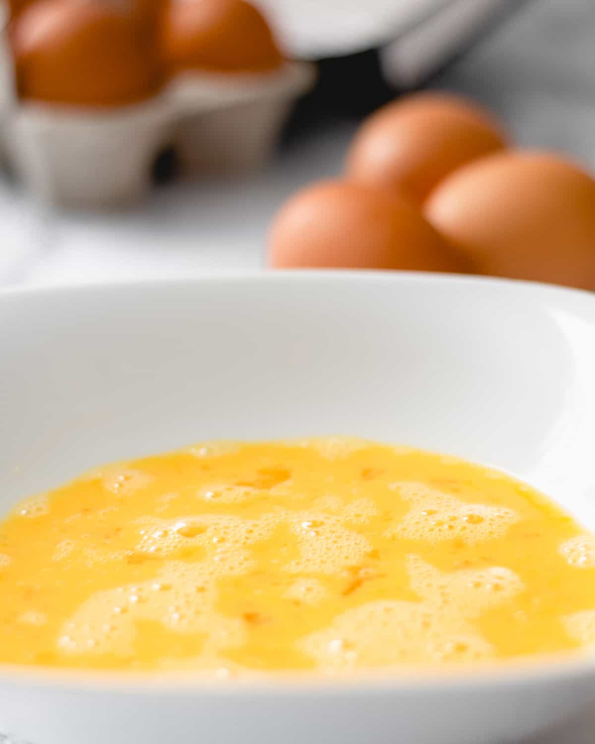 A bowl of beaten eggs, with whole eggs behind it.