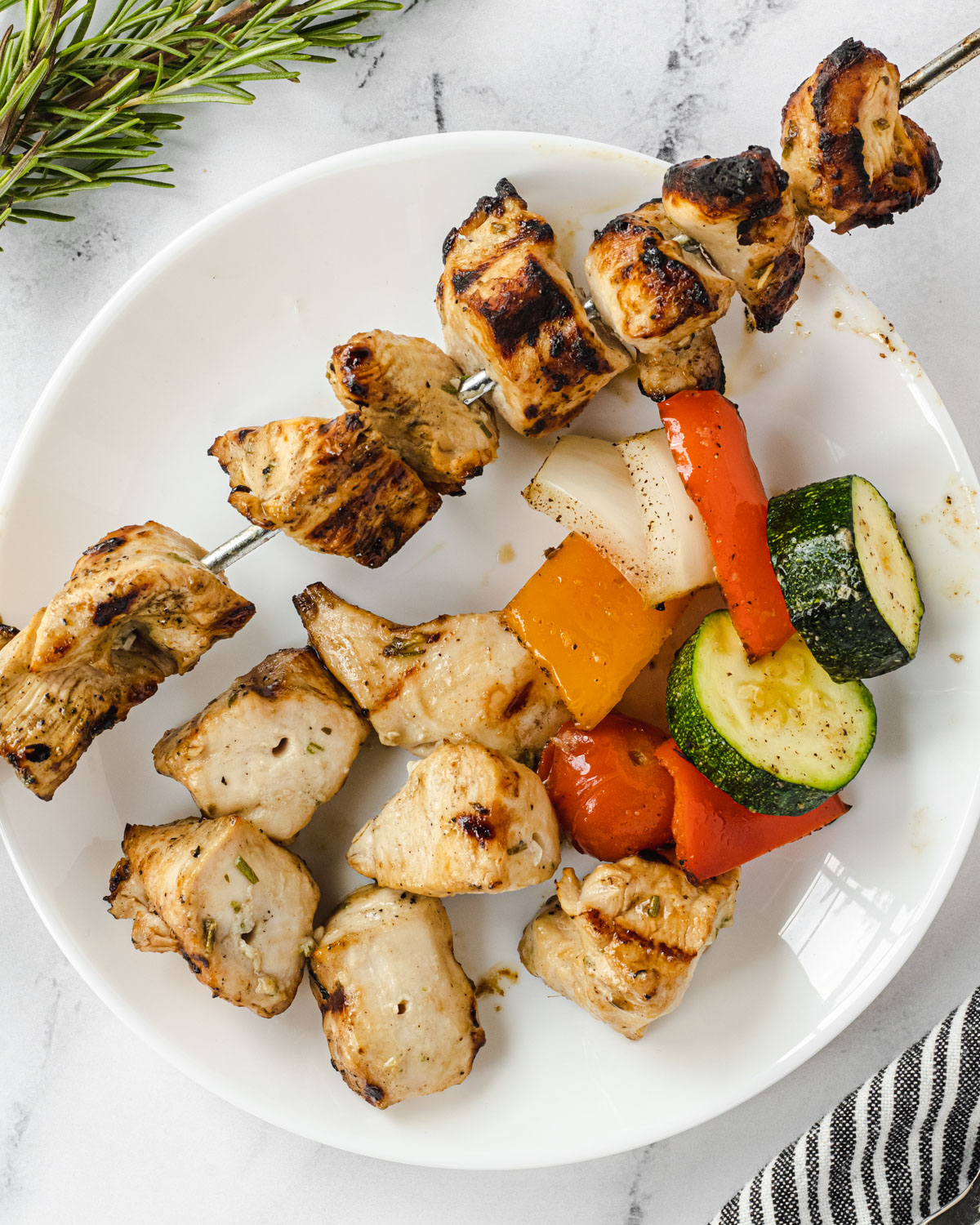 A skewer of rosemary ranch chicken on a white plate, with some pieces of chicken and grilled vegetables on the plate.