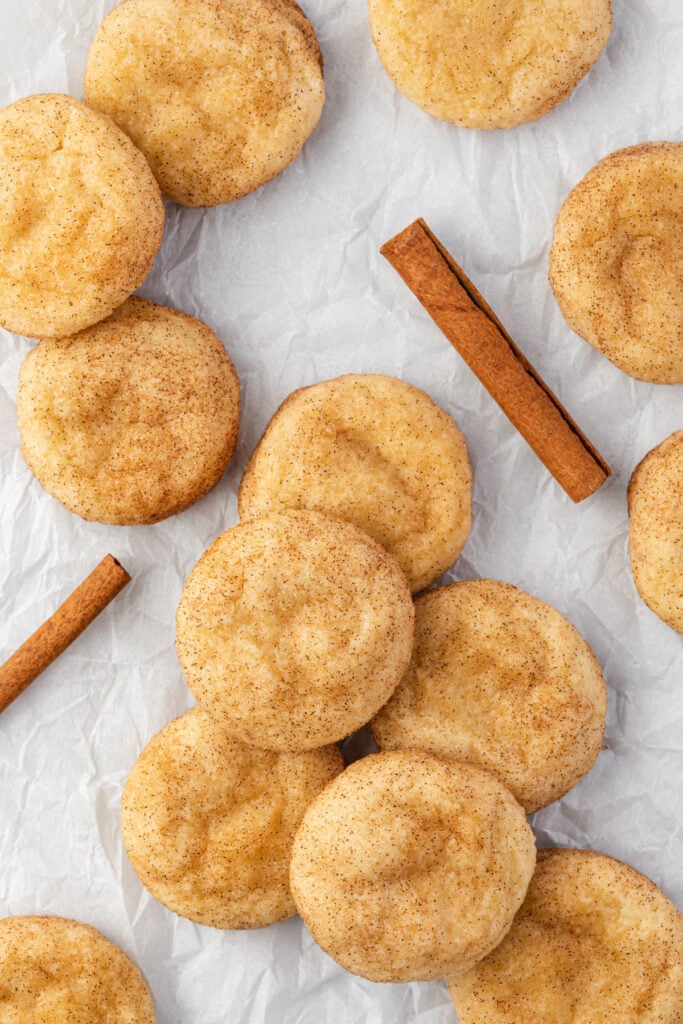 A bunch of snickerdoodle cookies clustered on a sheet of parchment paper with 2 cinnamon sticks between them.