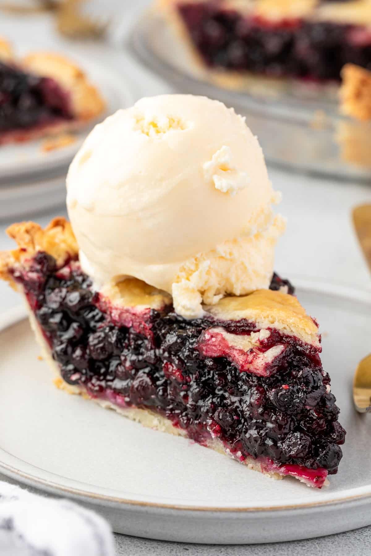 A slice of pie topped with a big scoop of vanilla ice cream, with another slice of pie and the whole pie in background.
