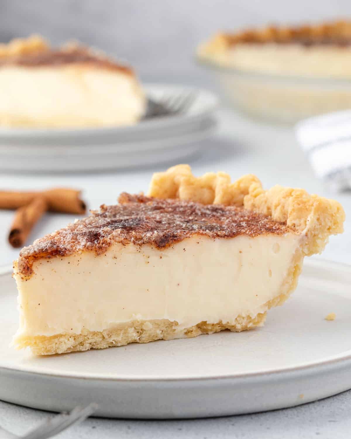 Side view of sugar cream pie slice on a plate, with 2 cinnamon sticks on counter behind it.