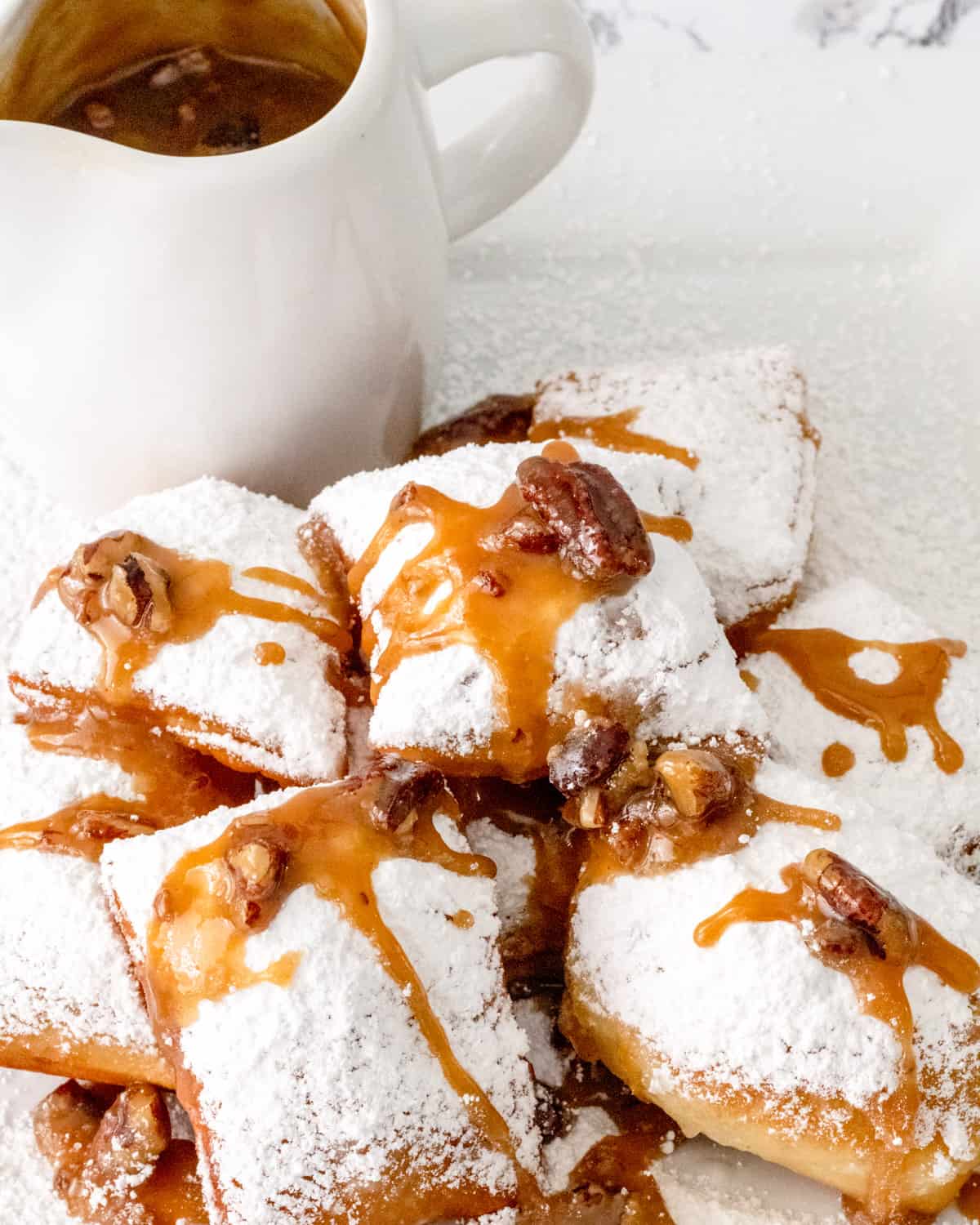 Plate of beignets covered in powdered sugar with pecan praline sauce drizzled over. Small pitcher of praline sauce behind.