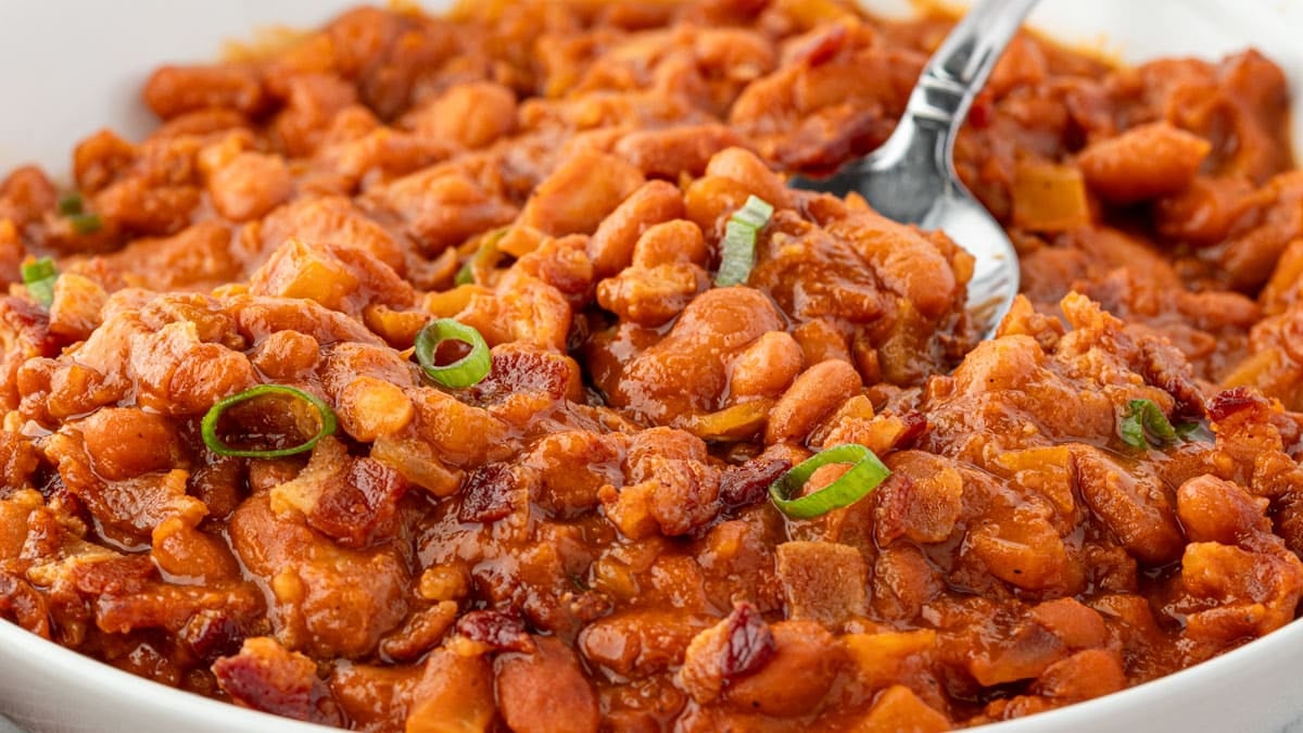 A close-up of baked beans in sauce, garnished with sliced green onions, with a spoon in the dish.