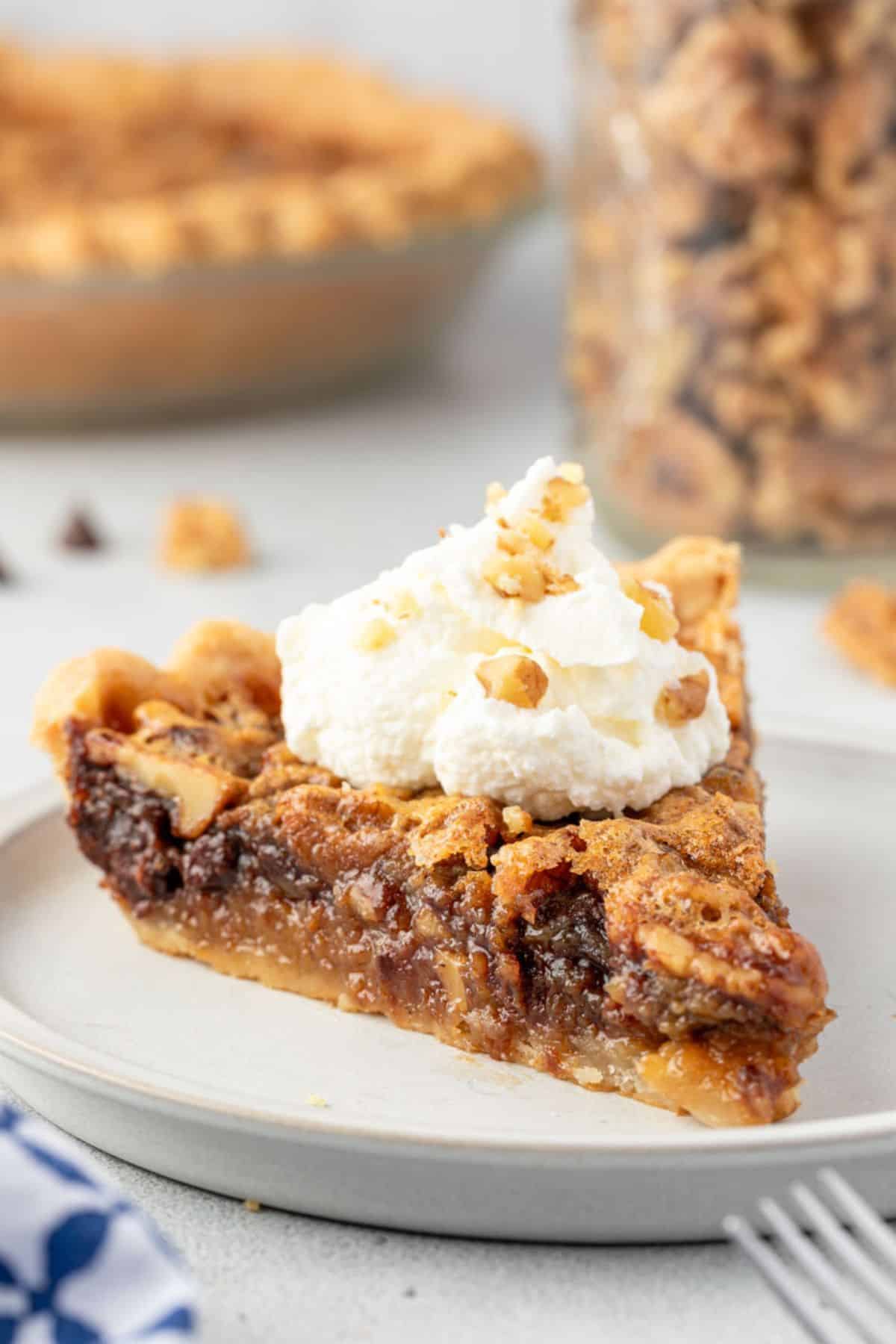 A slice of pie topped with whipped cream, with the whole pie and a jar of walnuts in background.