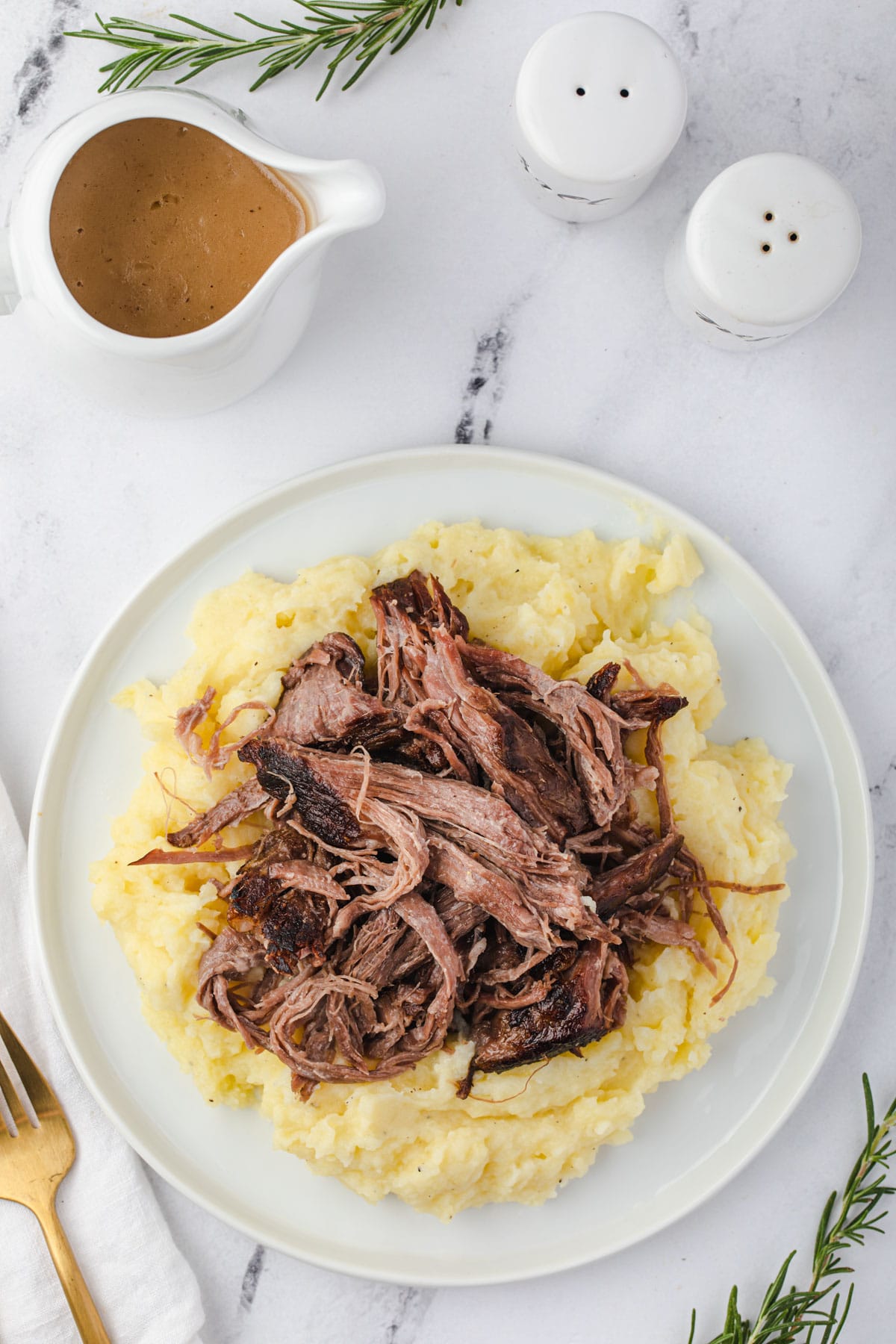 Shredded pot roast on a pile of mashed potatoes with small pitcher of gravy and salt and pepper shakers at top. 
