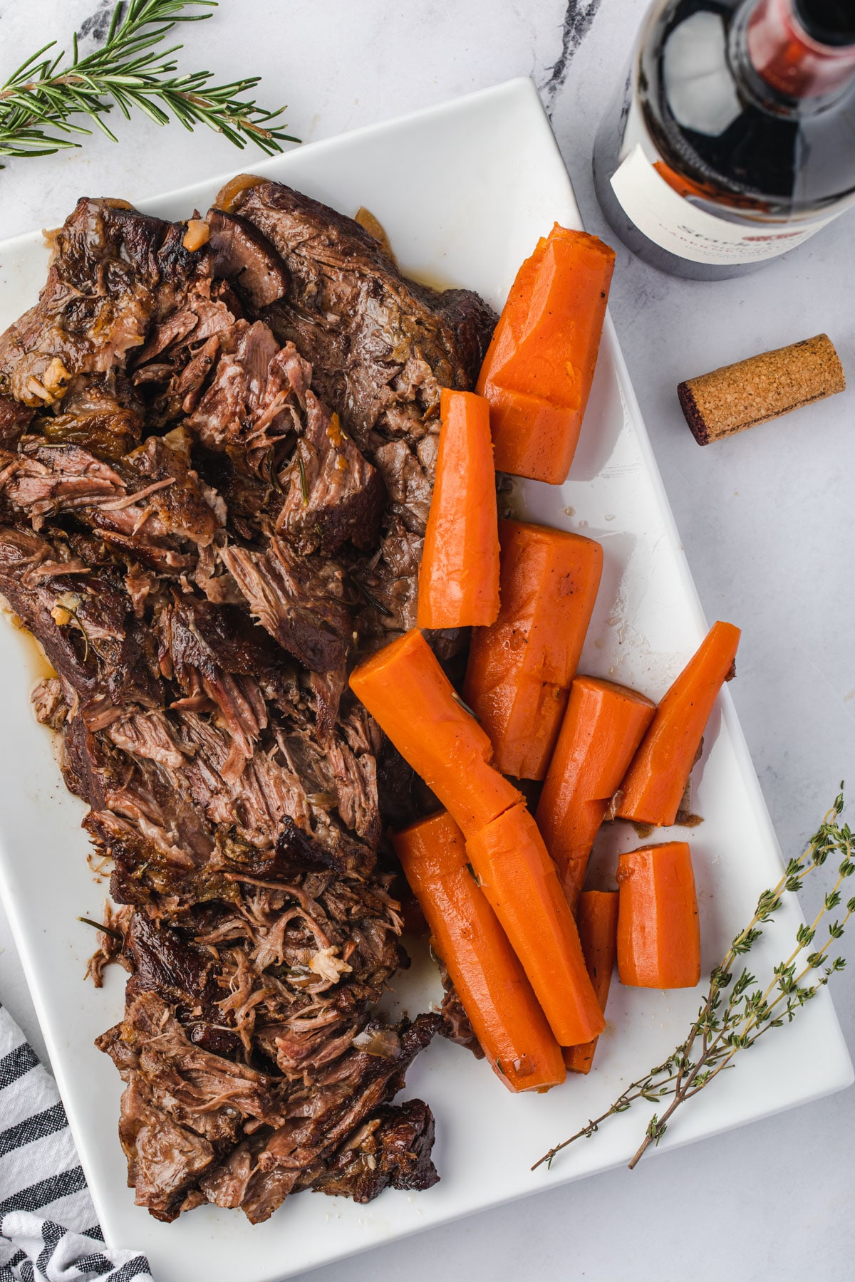 Shredded tender pot roast on a rectangular tray with roasted carrots and a bottle of red wine.