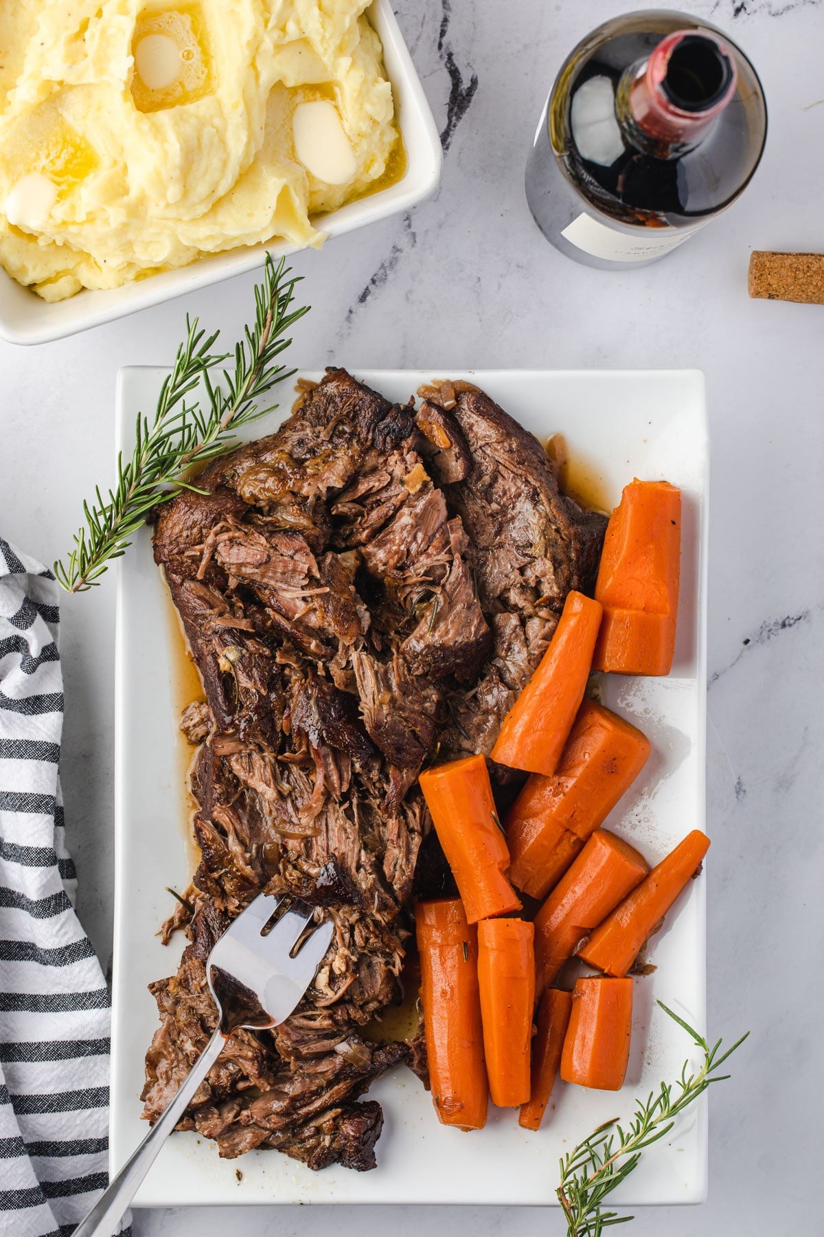 Shredded tender pot roast on a rectangular tray with roasted carrots and a bottle of red wine. and mashed potatoes in corners.