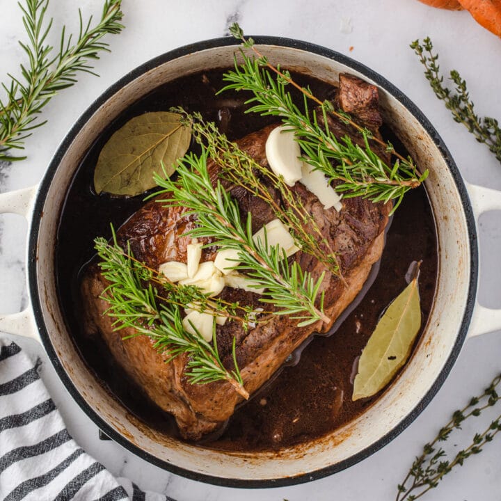 Pot roast in a dutch oven topped with garlic cloves, rosemary, and thyme sprigs.