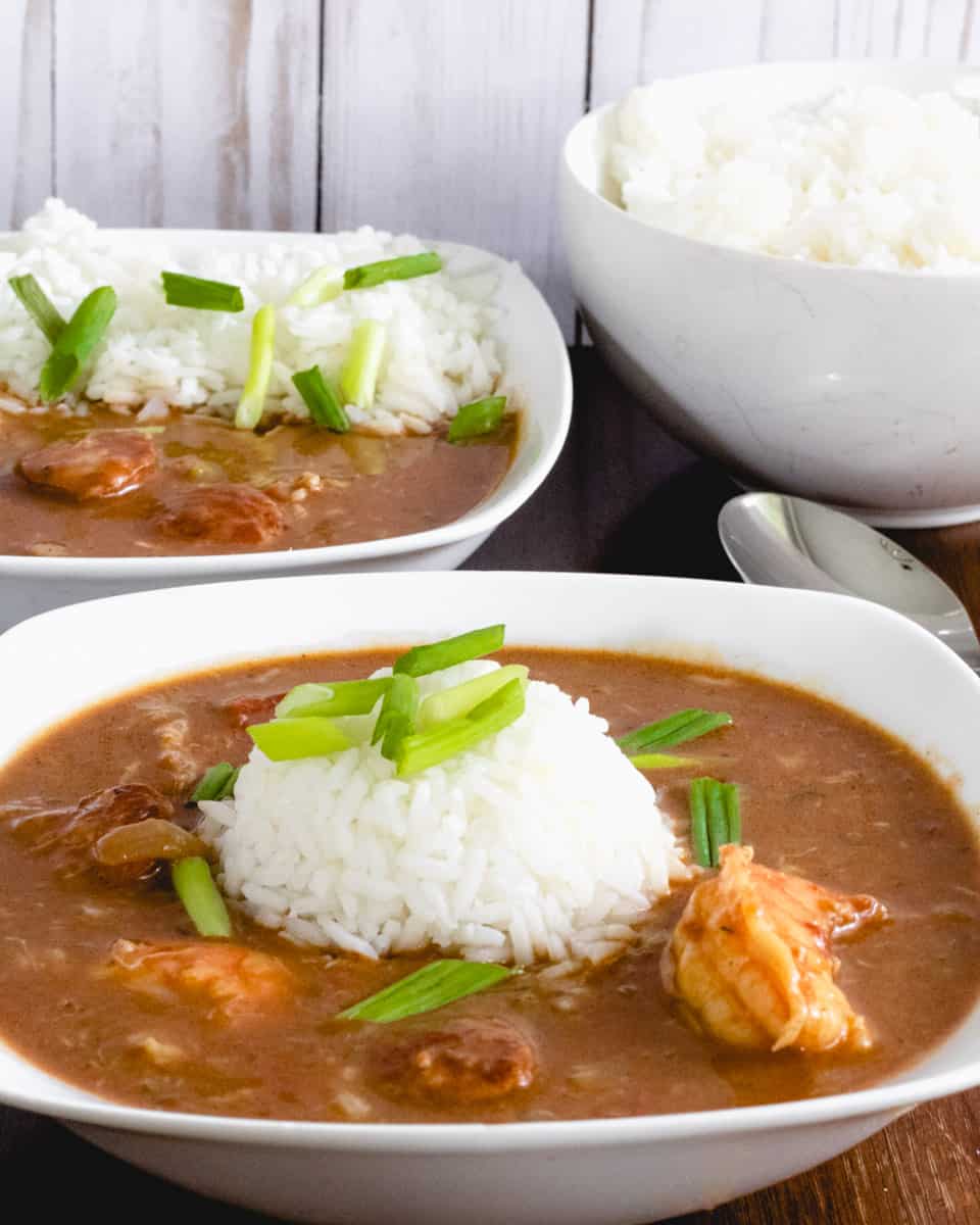 Bowl of gumbo with scoop of rice in the middle. Second bowl of gumbo and a bowl of rice behind.