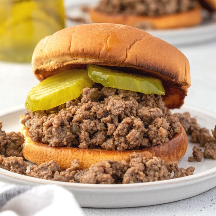 An individual loose meatsandwich on a plate with pickles and a mess of meat crumbles.