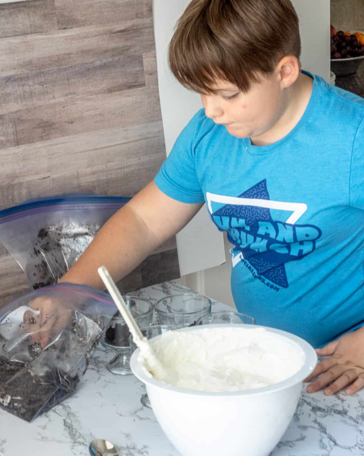 Boy with a blue shirt getting Oreo crumbs from a bag to put into the parfait bowls. A large bowl of whipped filling is next to him.