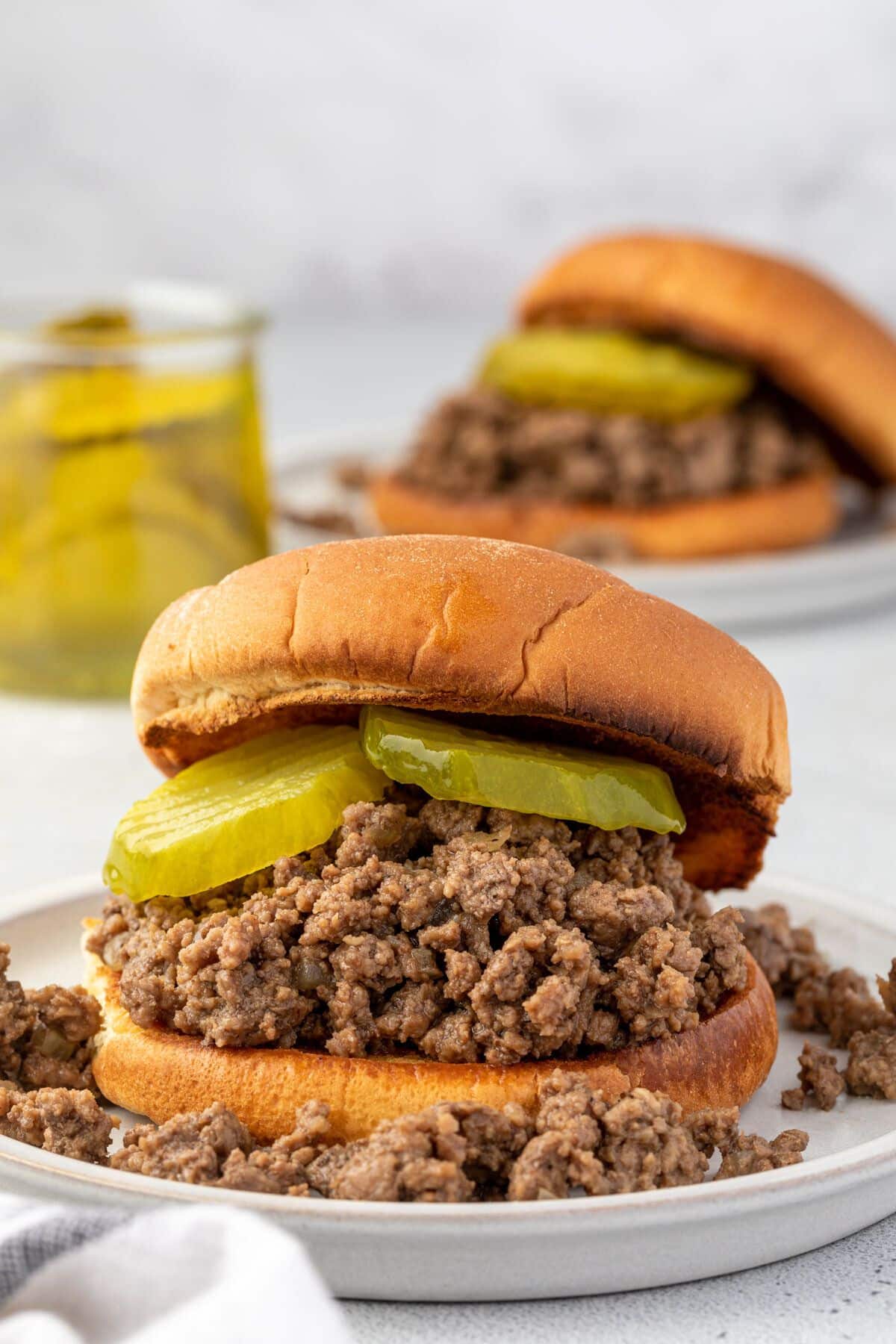 A hamburger bun filled with meat crumbles that are spilling out on a small plate, 2 pickle rings, topped with a bun.