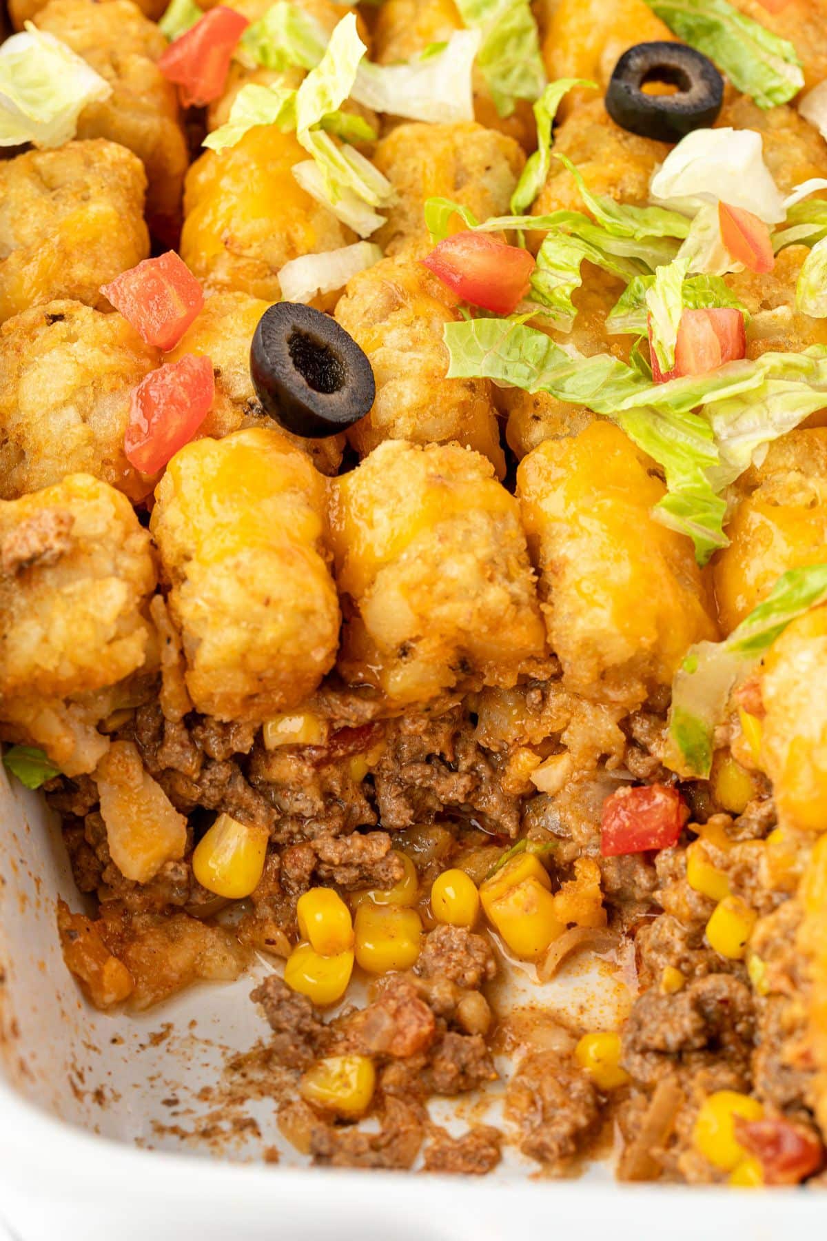 Close-up of tater tot casserole with ground beef, corn, melted cheese, lettuce, tomato, and black olive slices.