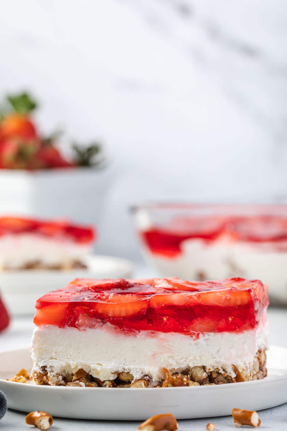 Piece of layered strawberry pretzel salad on a plate with the pan of jello salad and a basket of strawberries in the background.