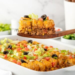 A wooden spatula lifts a cheesy taco casserole topped with lettuce, tomato, and olives from a white baking dish.