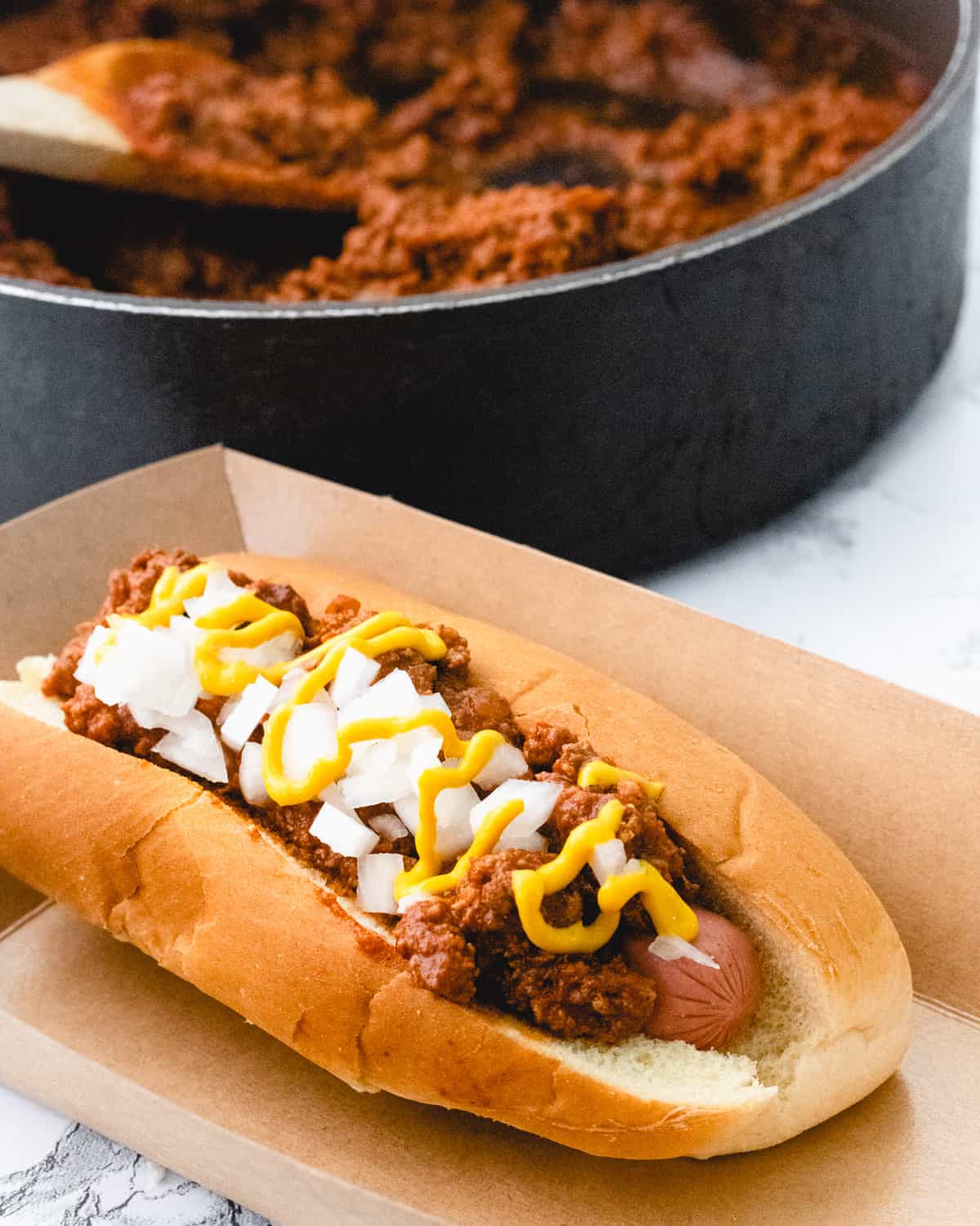 A coney dog in a cardboard holder with pan of chili sauce behind it.