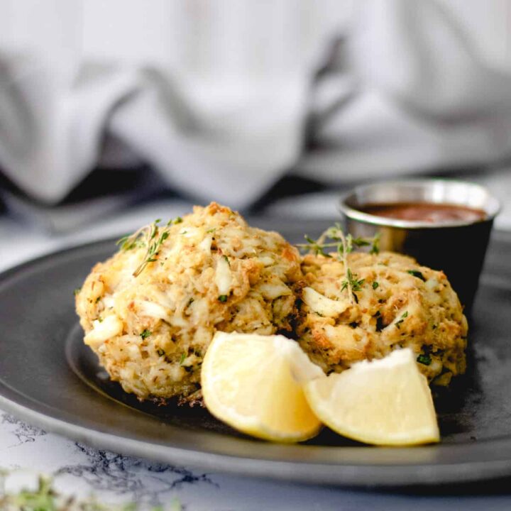 Two crab cakes on a dark grey plate, 2 lemon wedges in front and a ramekin of cocktail sauce in the back.