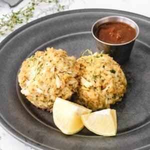 Two crab cakes, on a plate with lemon wedges and cocktail sauce.