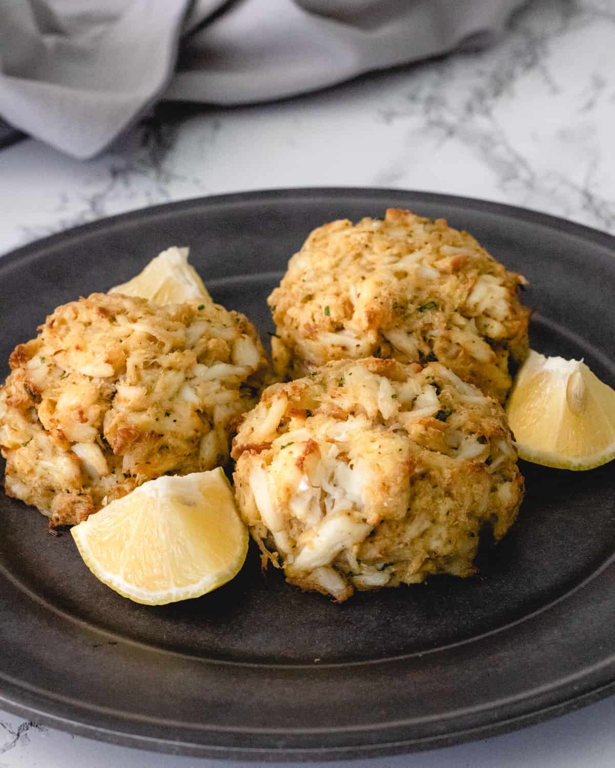 3 crab cakes on a dark grey plate, with lemon wedges in between them.