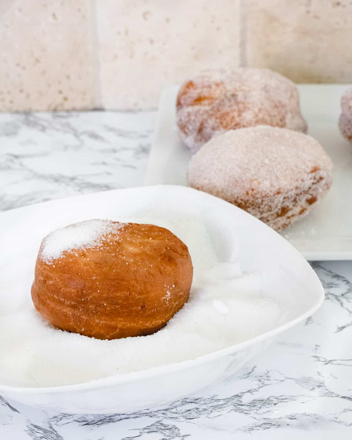 A paczki in a bowl of sugar. Paczki already covered with sugar are on a plate in the background.
