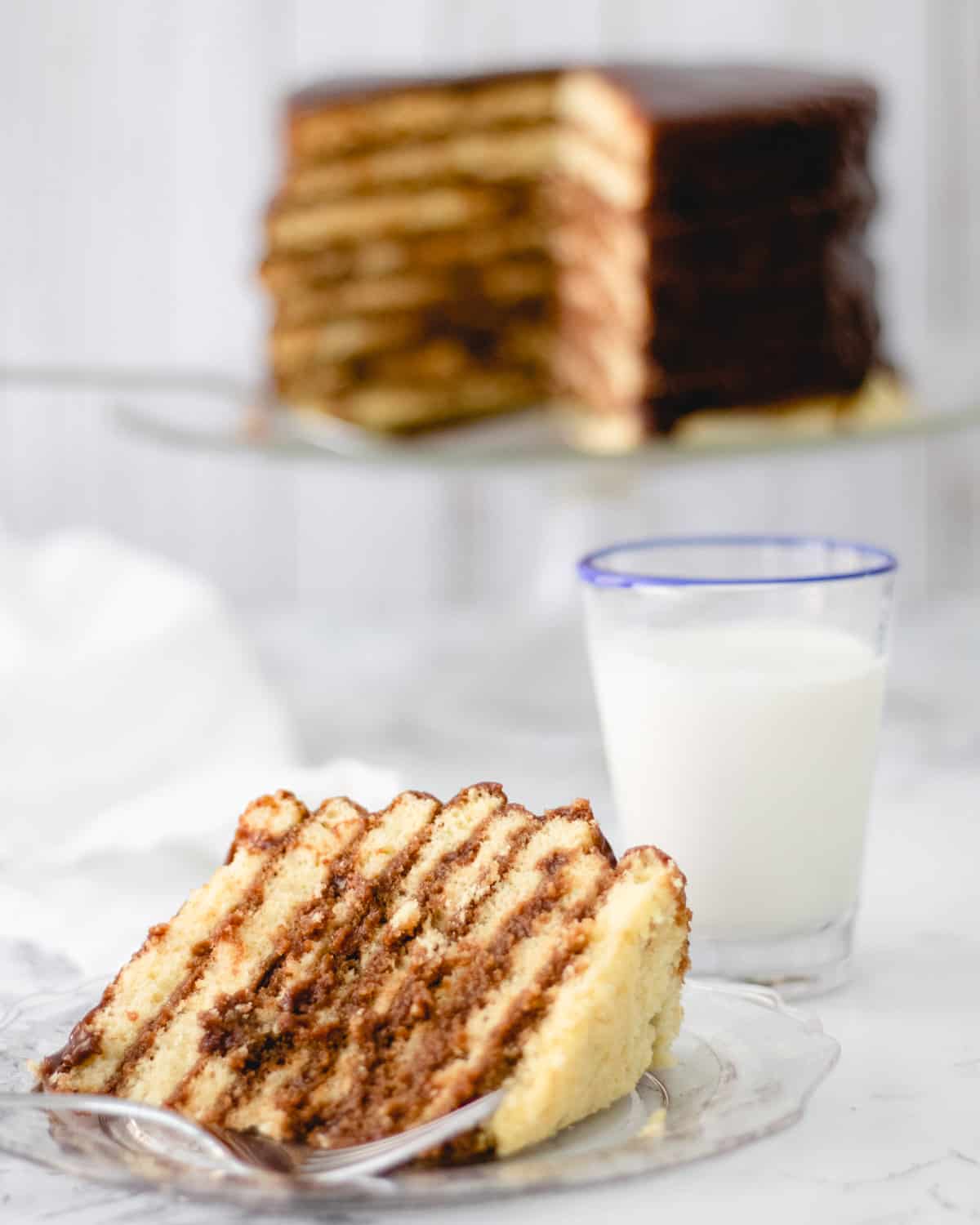 Slice of Smith Island Cake. Glass of milk and whole cake in background.