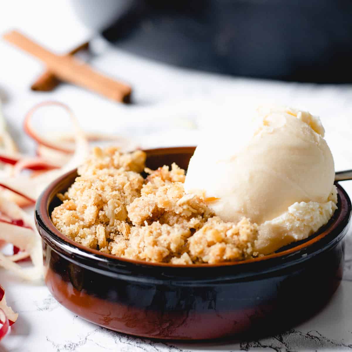 Individual serving of skillet apple crisp with ice cream. Apple peel ribbons and cinnamon sticks on the left.