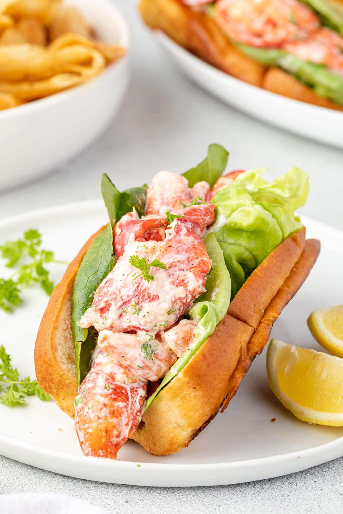 Mayo coated lobster pieces in a bun lined with lettuce, on a plate with lemon wedges.