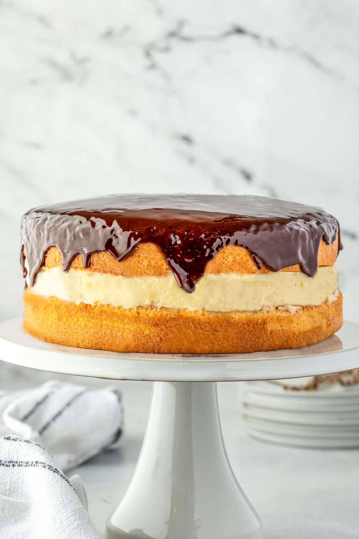 A layered cake on a cake stand, with cream filling and chocolate ganache dripping down the sides.