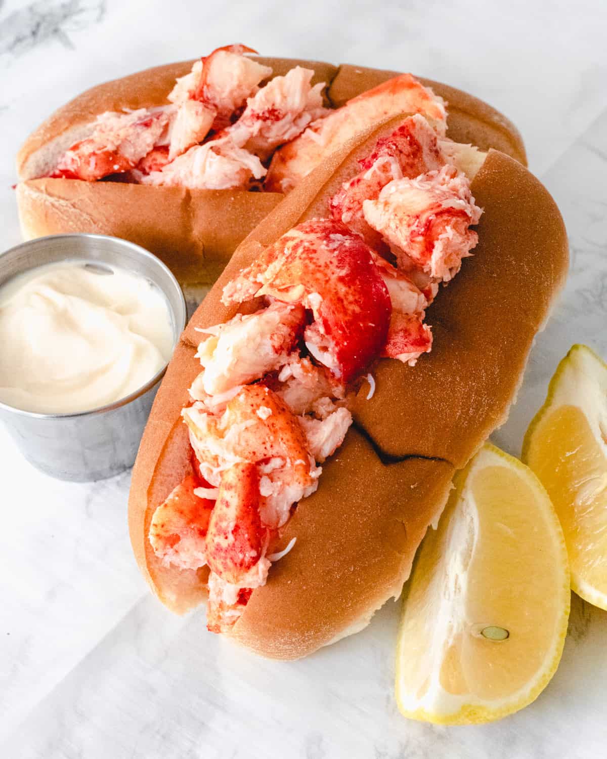 Two lobster rolls with a ramekin of mayo on the left and 2 lemon wedges on the right.