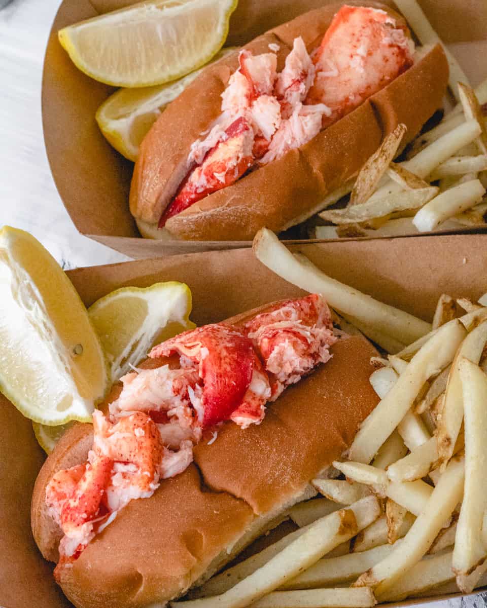 Two lobster rolls in cardboard trays. Each with lemon wedges and French fries.