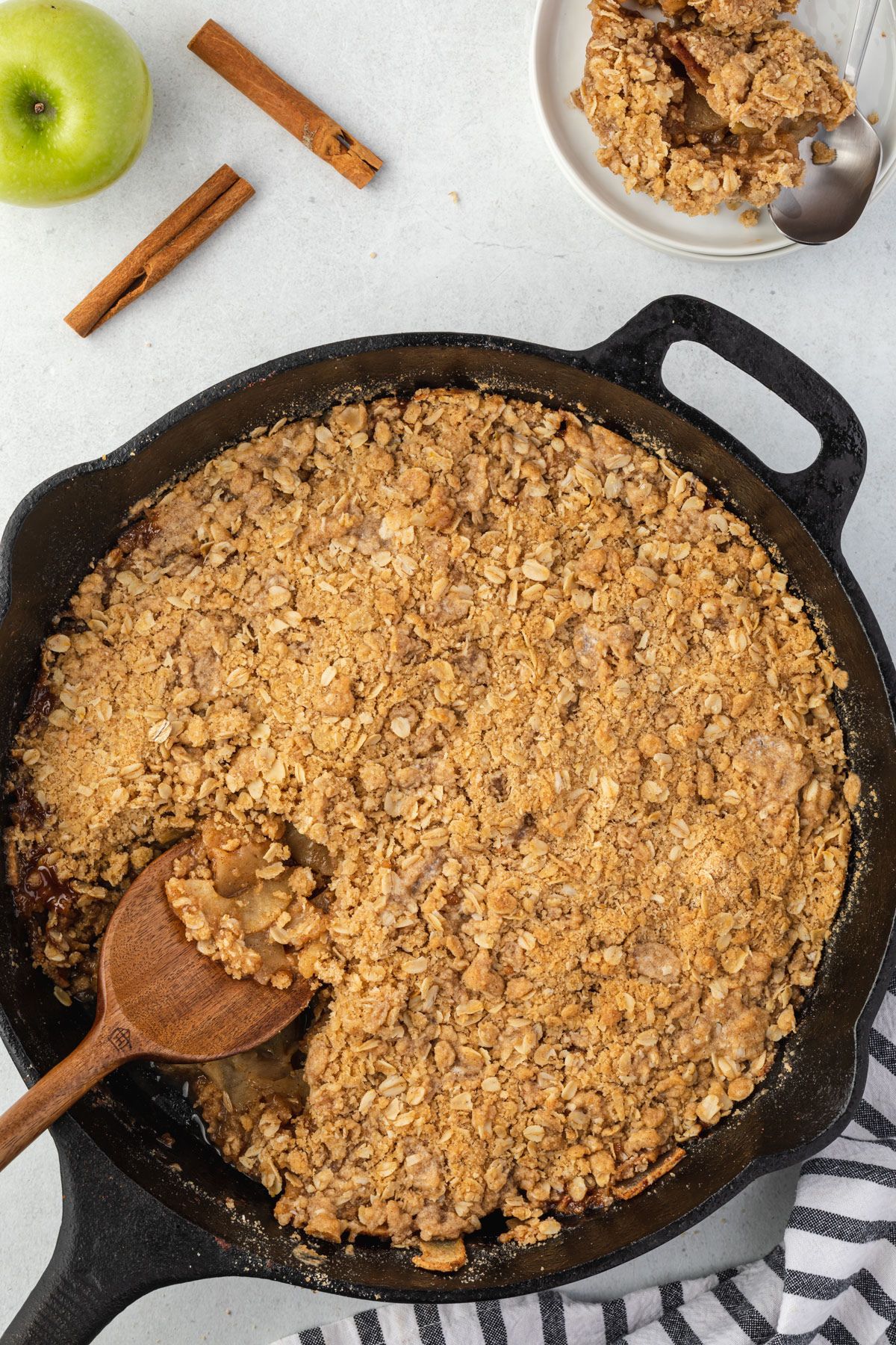 Baked apples slices with a crumbling oat topping baked in a black cast iron skillet with a wooden spoon scooping out some apple crisp.