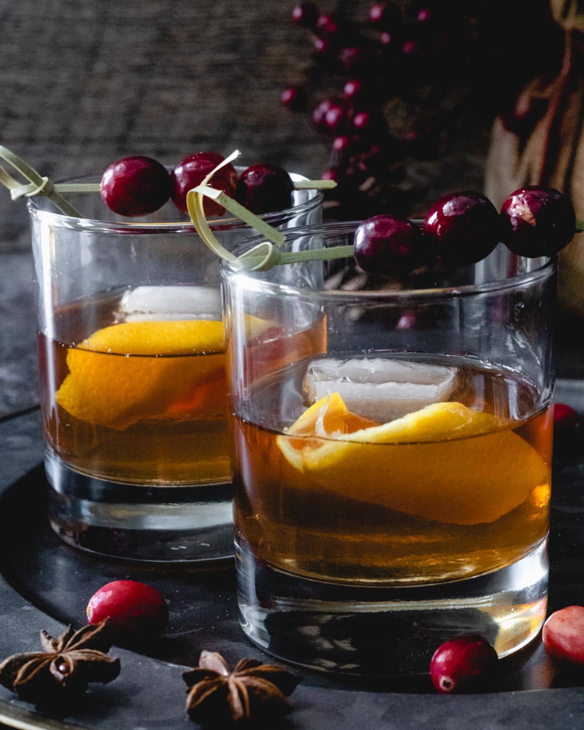 Two cranberry old fashioneds on a tray, with orange peel in glass and cocktail picks with cranberries.