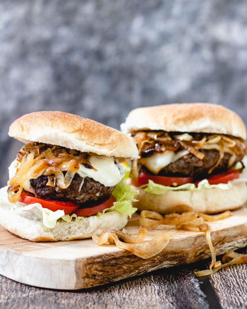 Two elk burgers on a wooden board with caramelized onions and cheese.