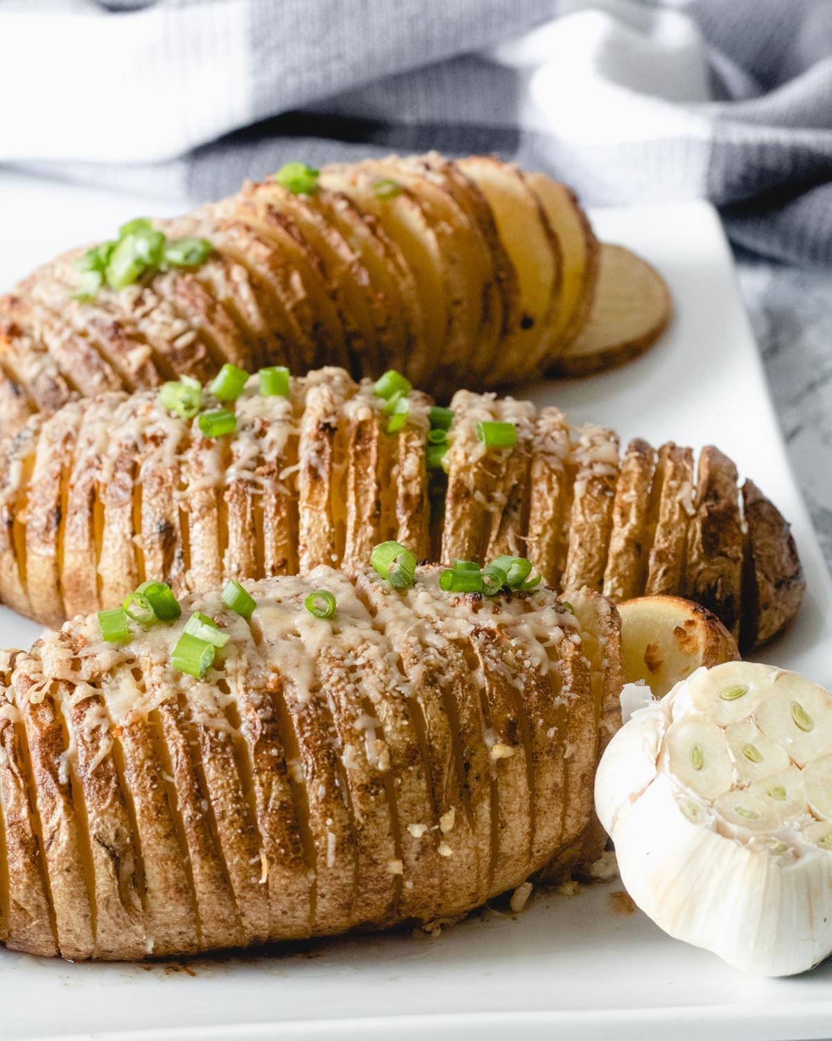 Garlic Parmesan Hasselback Potatoes topped with green onions, on a plater with a head of garlic.