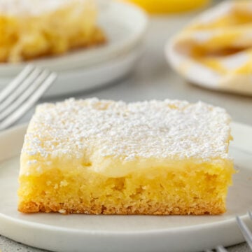 A square piece of gooey lemon butter cake topped with powdered sugar on a white plate.