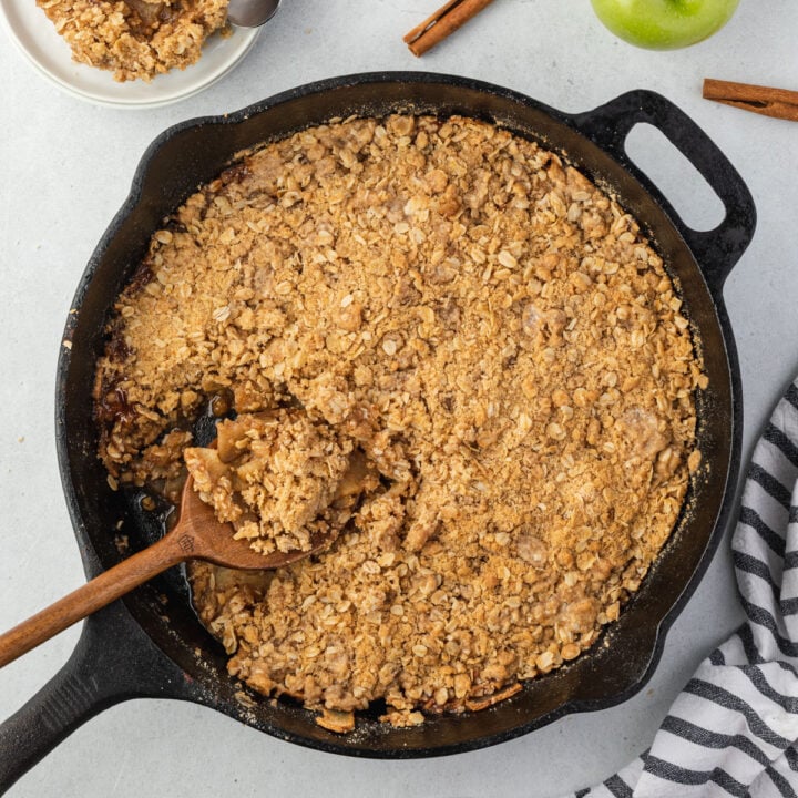 A wooden spoon scooping out a serving of apple crisp that is in a cast iron skillet.