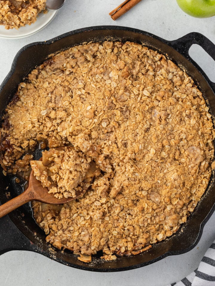 A wooden spoon scooping out a serving of apple crisp that is in a cast iron skillet.