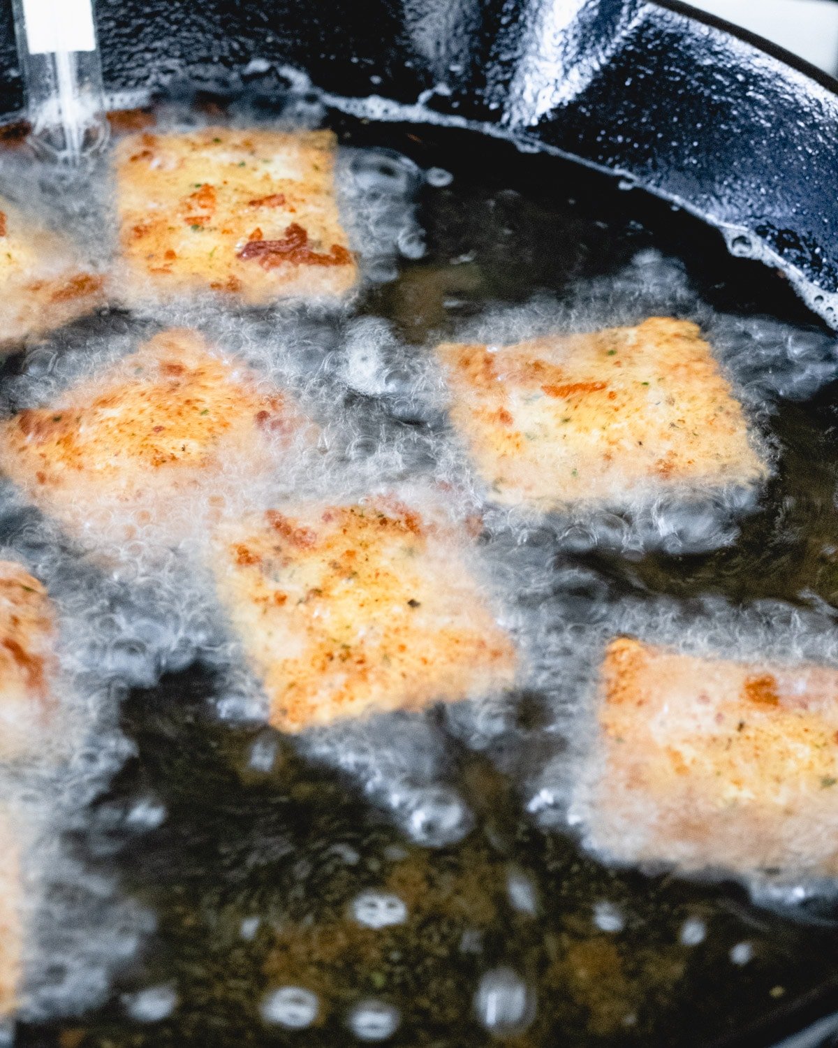 Breaded ravioli being deep fried in a cast iron skillet.