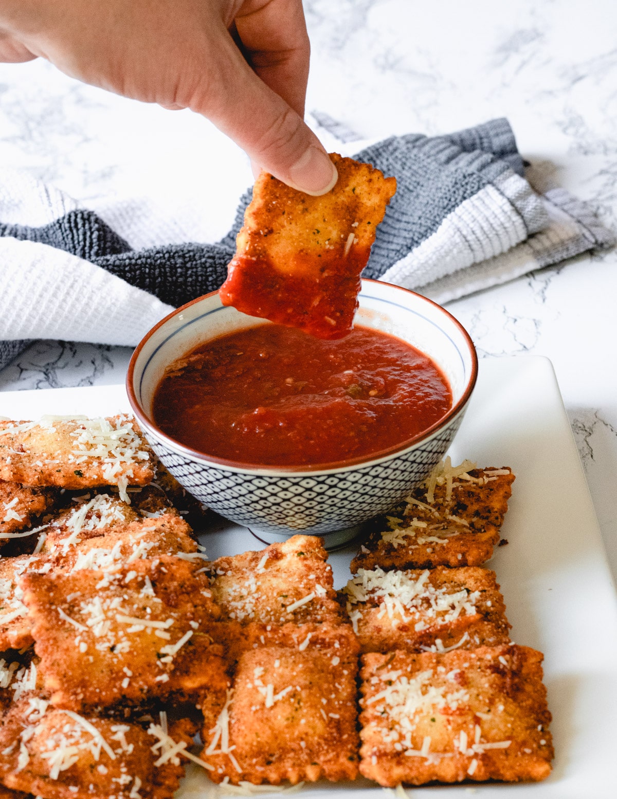 A hand dipping toasted ravioli into a bowl of marinara sauce, with a platter of toasted ravioli.