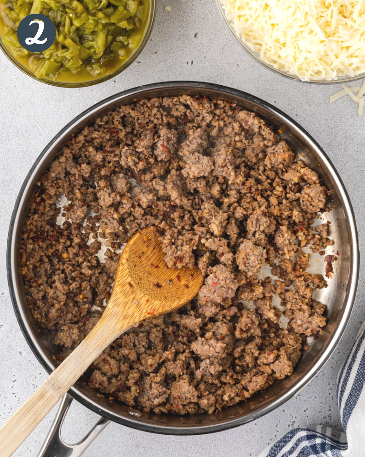 Ground sausage in a pan with a wooden spoon, and green chiles and cheese above.