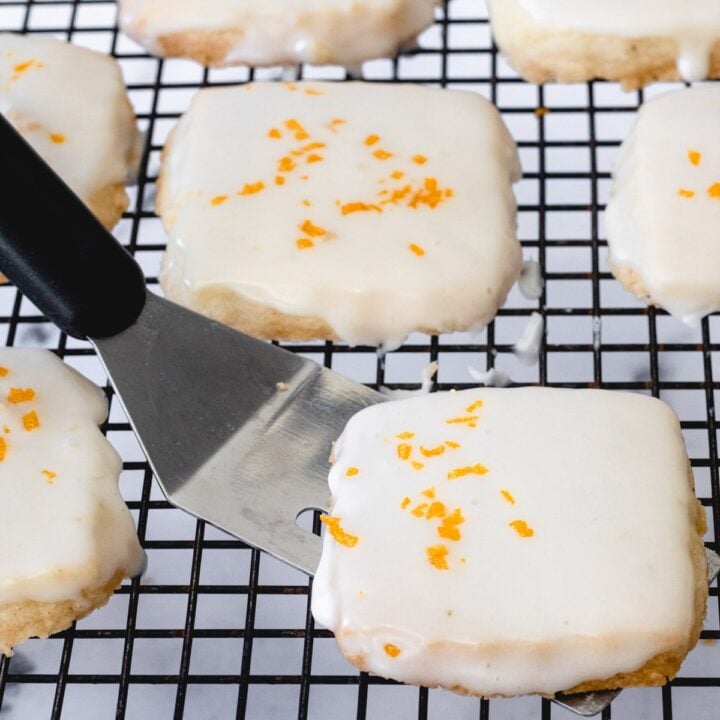 Square cardamom shortbread cookies with a citrus glaze, on a cooling rack with a mini spatula under one cookie.