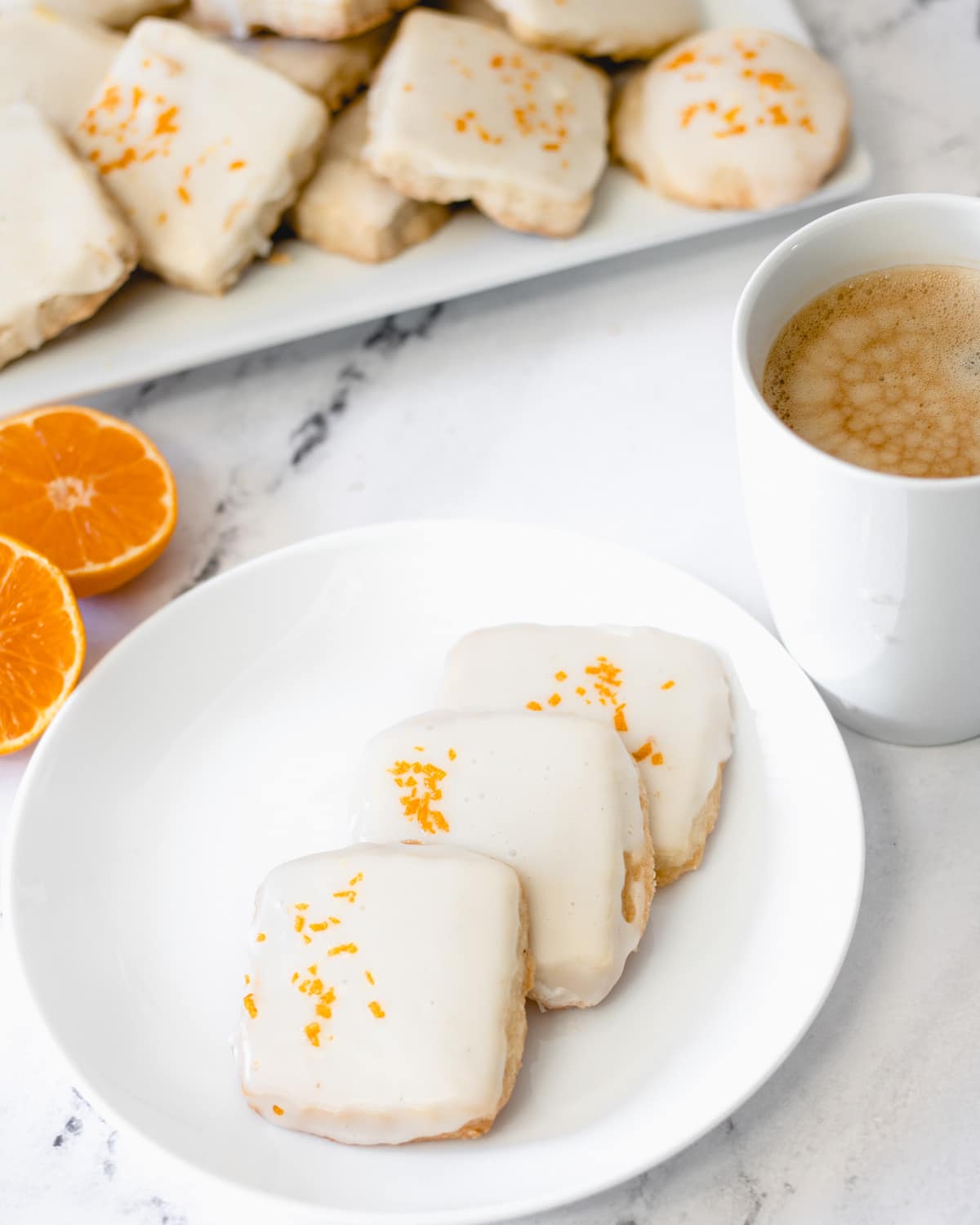 A round plate with 3 cardamom shortbread cookies, a cup of coffee top right, and top left is a plate of cookies and a cut orange.