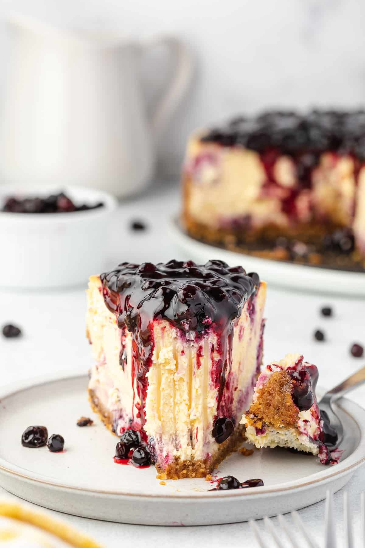 Slice of cheesecake on a plate with bite on a fork, whole cheesecake and bowl of huckleberries in background.