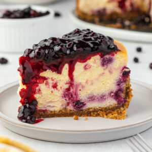 1 slice of cheesecake on a plate, huckleberry sauce dripping and huckleberries scattered behind.