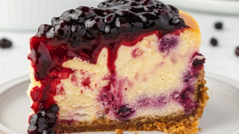 1 slice of cheesecake on a plate, huckleberry sauce dripping and huckleberries scattered behind.