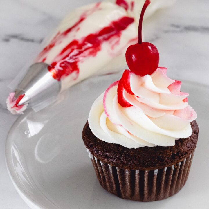 Chocolate cupcake with red and white swirl frosting, a red cherry on top, and next to it is a piping bag with white frosting and a red stripe.