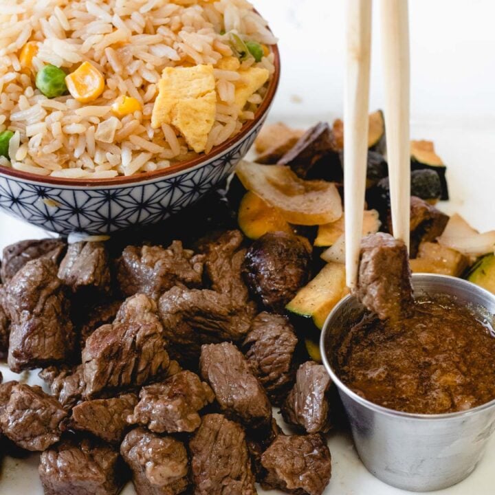 Plate of steak pieces, veggies, and fried rice with chop sticks dipping a piece of beef into ginger sauce.