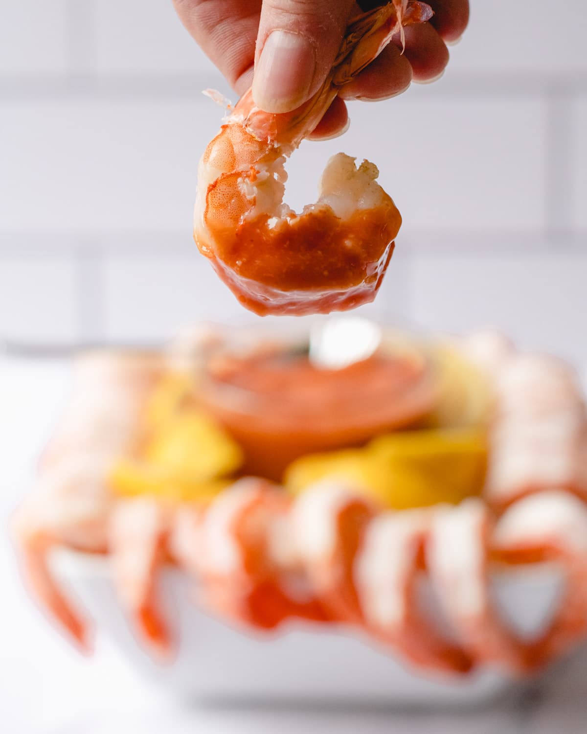 Close up of poached shrimp dipped in cocktail sauce, bowl of shrimp cocktail in background.